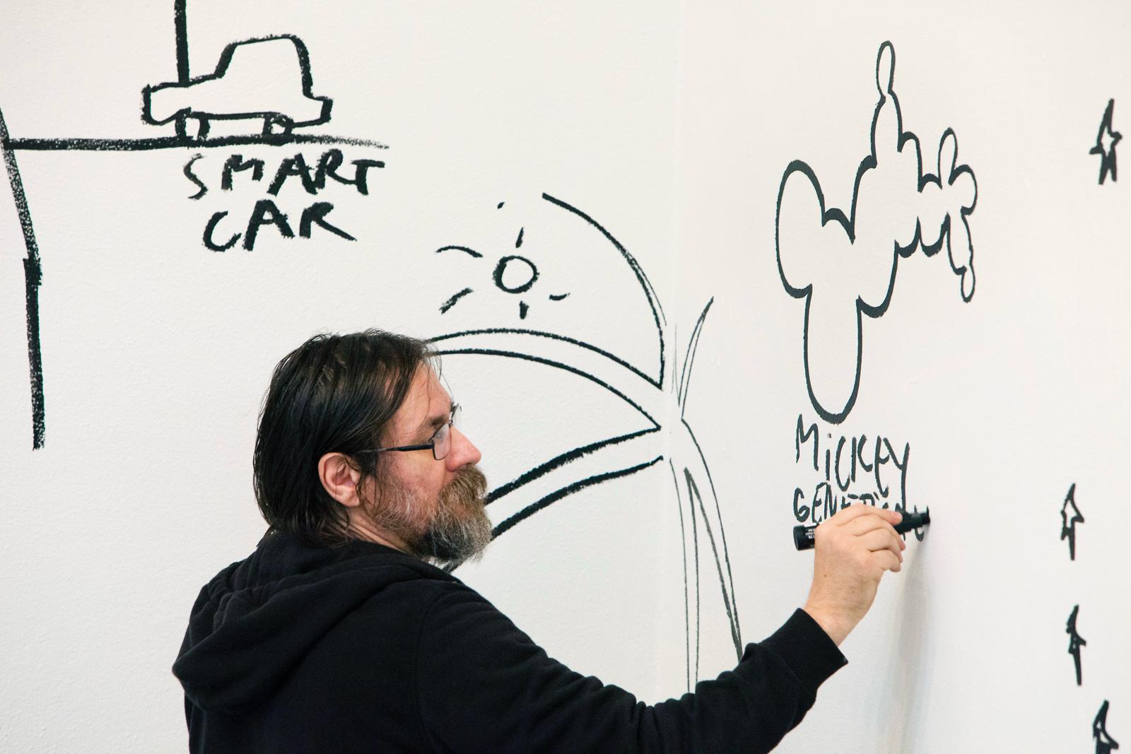 Der Künstler Dan Perjovschi, ein weißer Mann mittleren Alters mit mittellangen Haaren und Bart malt mit einem schwarzen Marker cartoon-artige Kunstwerke auf eine weiße Wand