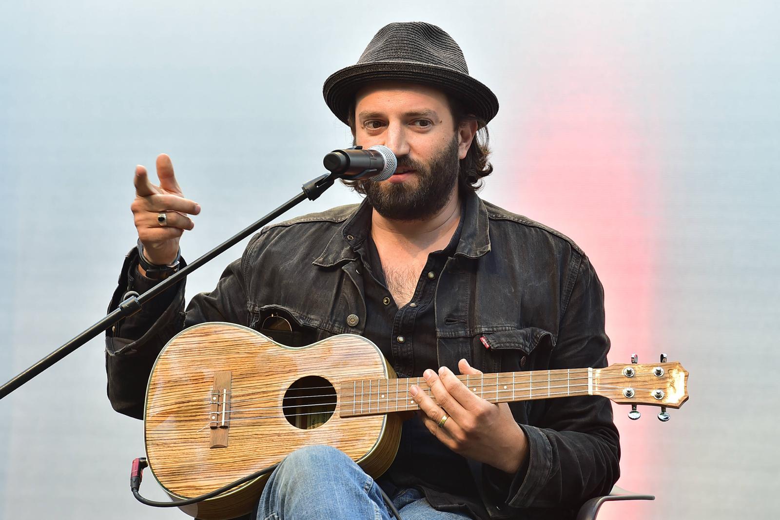 Der Musiker Daniel Kahn, ein junger weißer Mann mit scharzen, mittellangen Haaren, Bart und einem schwarzen Hut sitzt mit einer Ukulele auf dem Schoß vor einem Mikrofon